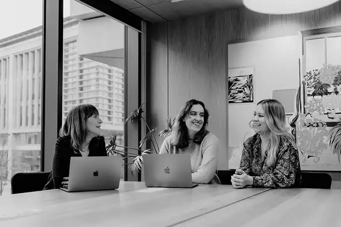 Black and white photo of the famille team. They are sitting in a meeting room with views over the city. They have two laptops and are engaging with each other and smiling
