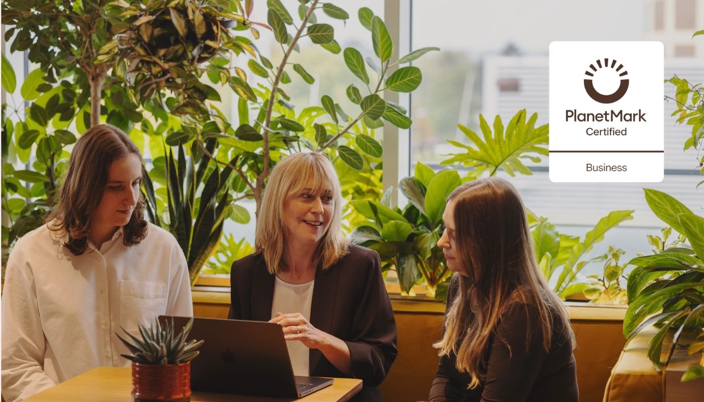 The team sit around a table with a laptop, surrounded by plants. A Planet Mark Certified Business logo is visible.